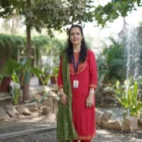 Female teacher in red traditional outfit standing outdoors in a garden area.